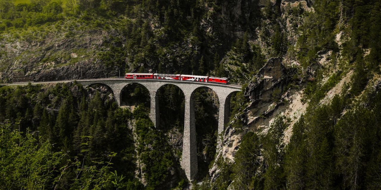 Red passenger train traveling on a stone viaduct, surrounded by lush green alpine scenery.
